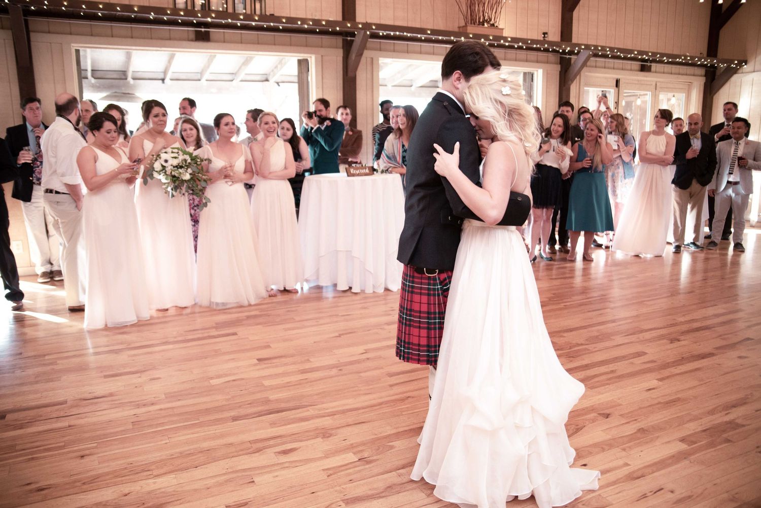 Bride and groom share first dance in rustic barn wedding venue with bridesmaids in pink gowns watching nearby.