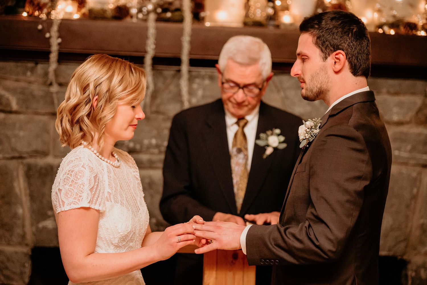Bride placing wedding ring on groom's finger during intimate ceremony with officiant before stone fireplace.