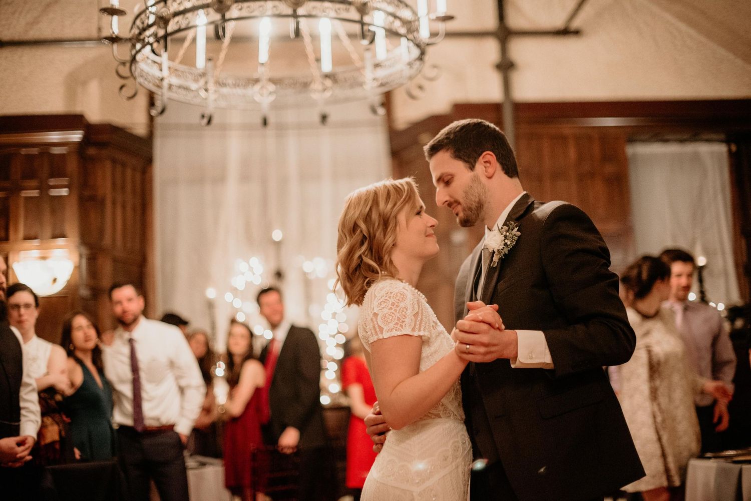 Couple shares their first dance at elegant wedding reception with chandelier and sparklers in background.