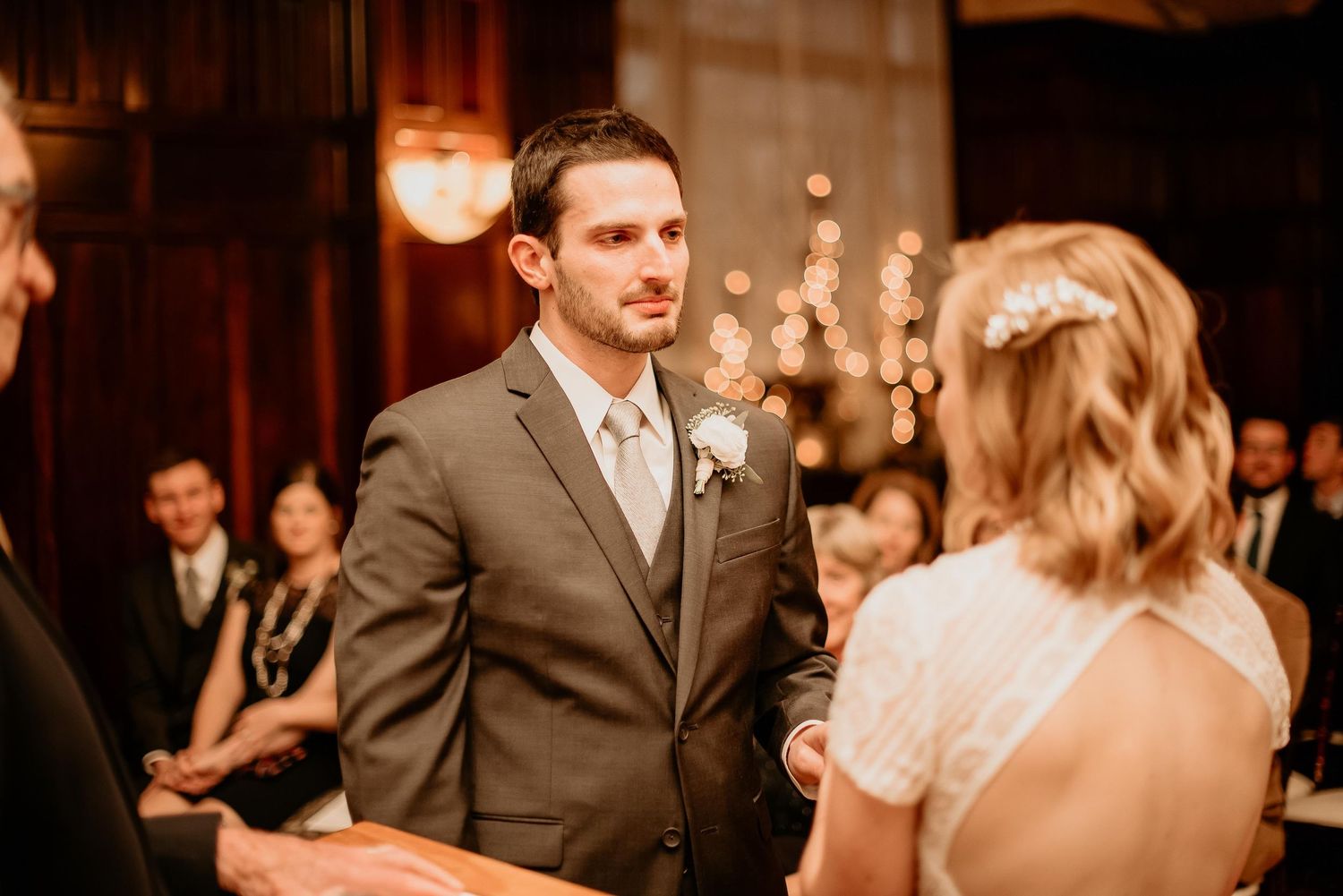 Groom in gray suit gazing lovingly at bride during indoor wedding ceremony with bokeh lights in background.