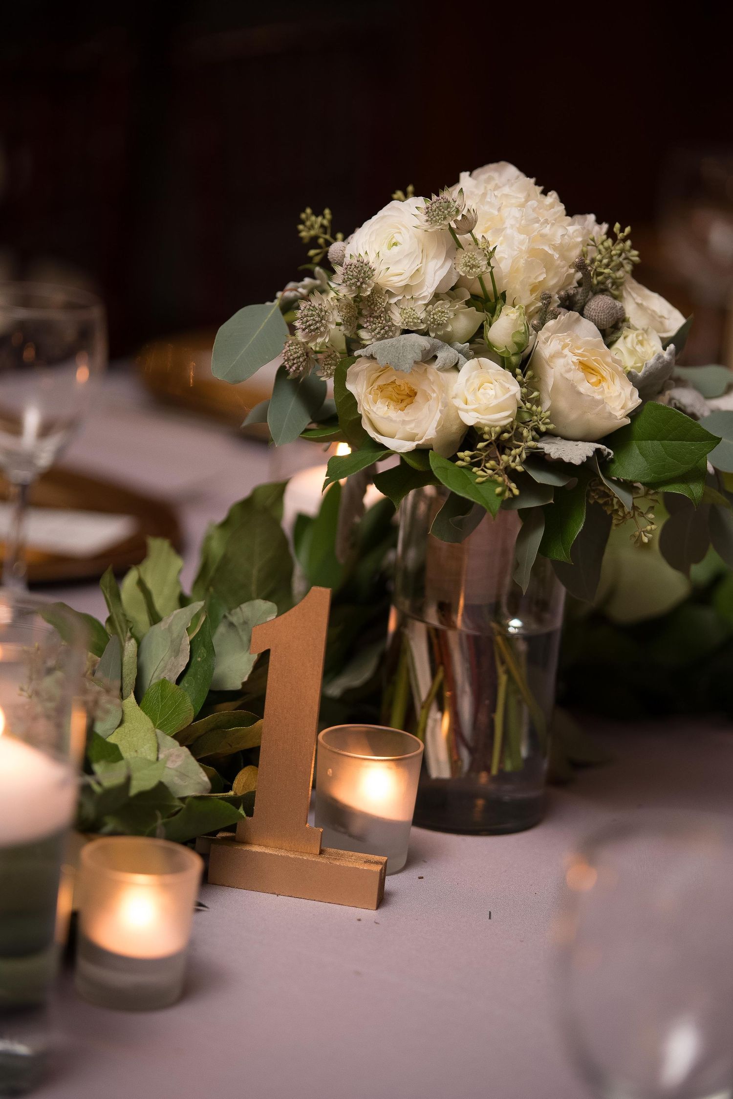 Elegant wedding table centerpiece with white roses, greenery, gold number one sign, and glowing votive candles.