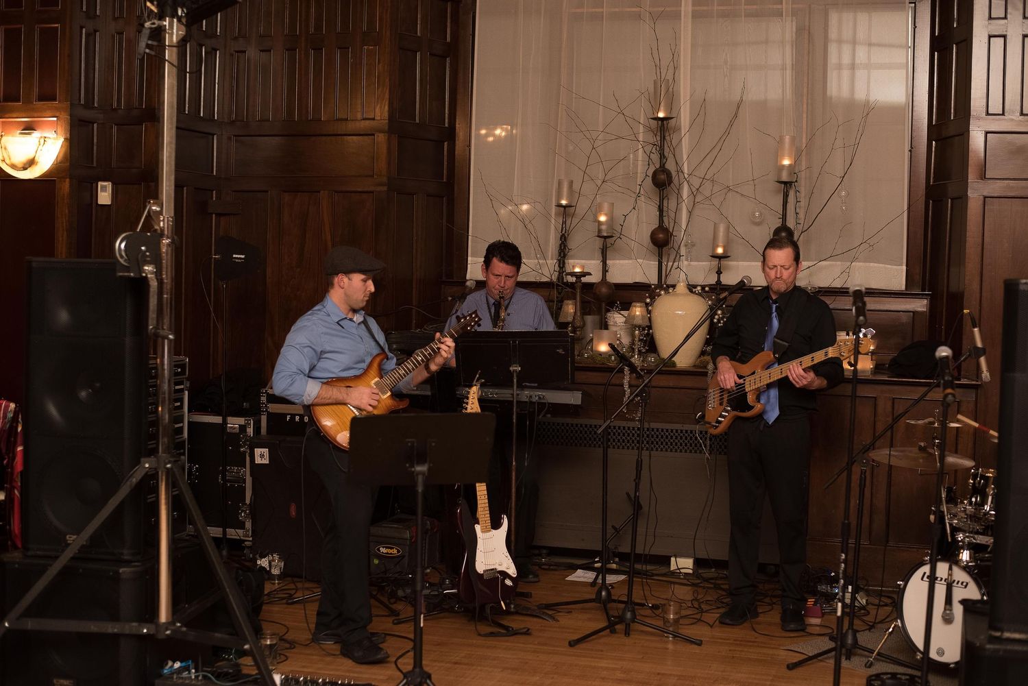 Three musicians performing on electric guitars in an elegant wood-paneled room with candlelit decor.