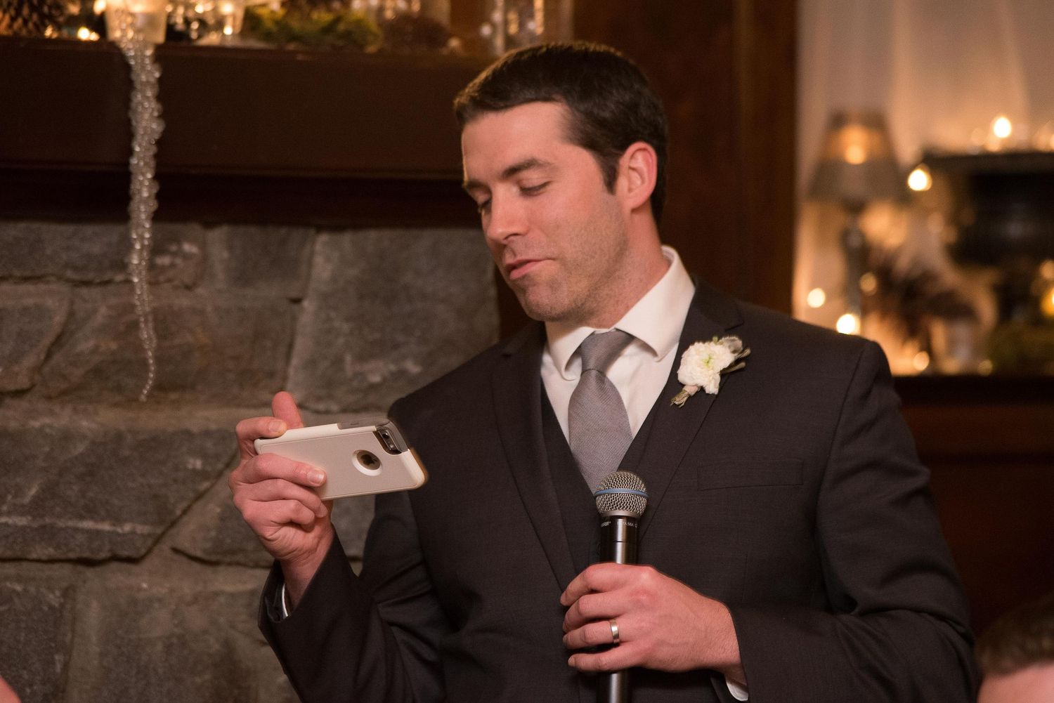 Groom in dark suit reading phone at wedding reception microphone, fireplace and candles in background.