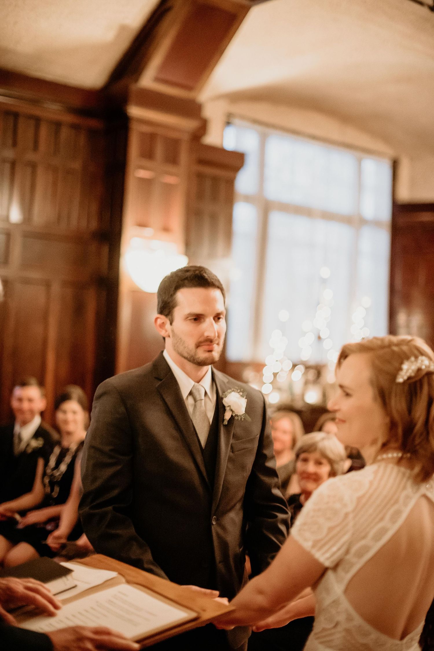 Groom in dark suit gazes at bride during indoor wedding ceremony in elegant wood-paneled room with guests seated behind.