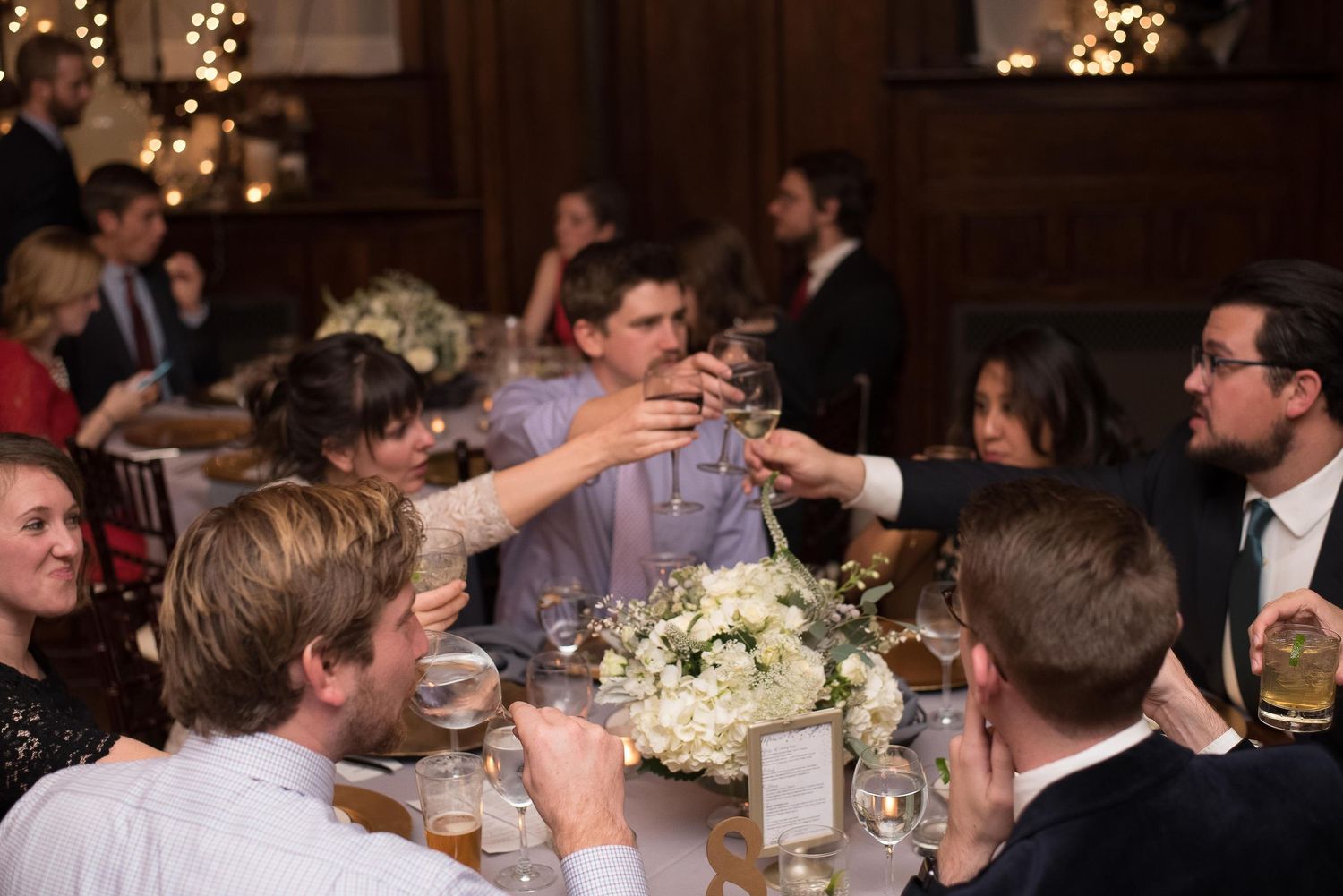 Wedding guests raising glasses in a toast at an elegant reception with white floral centerpieces and warm string lights.