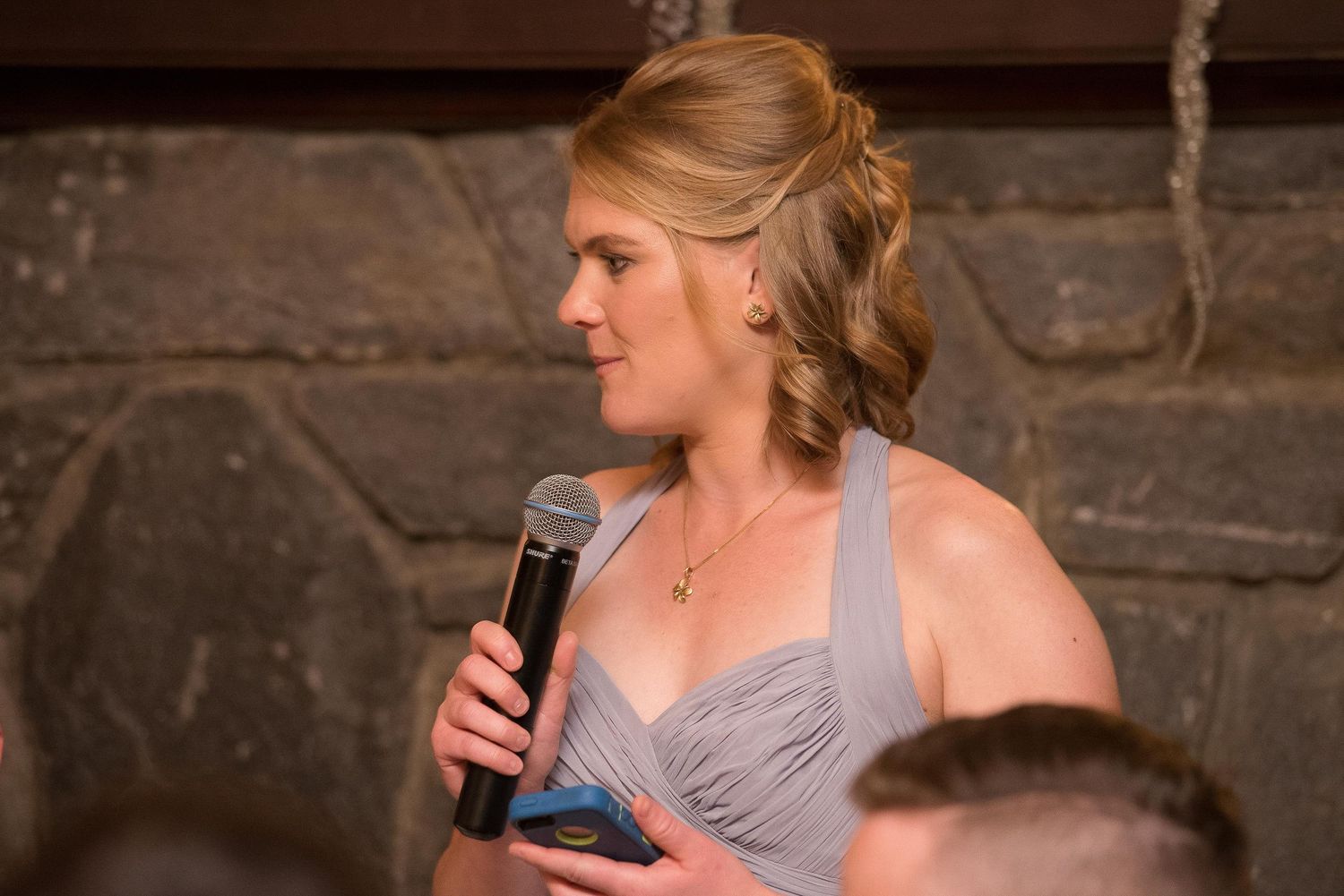 Woman in gray bridesmaid dress giving a speech at a wedding reception while holding a microphone.