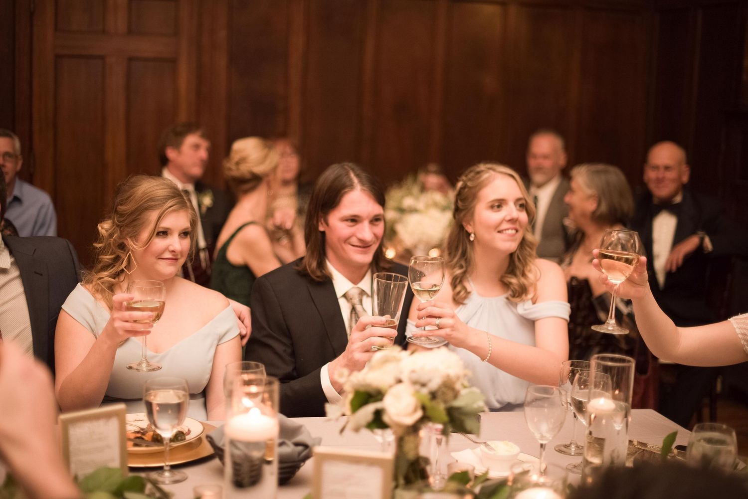Guests raising glasses in a toast at an elegant wedding reception with candlelit tables and floral centerpieces.