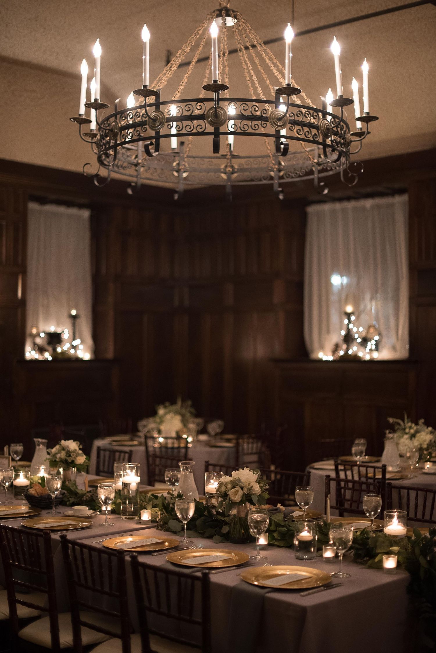 Elegant wedding reception with iron chandelier, candlelit tables, gold chargers, and white floral centerpieces in a dark wood room.