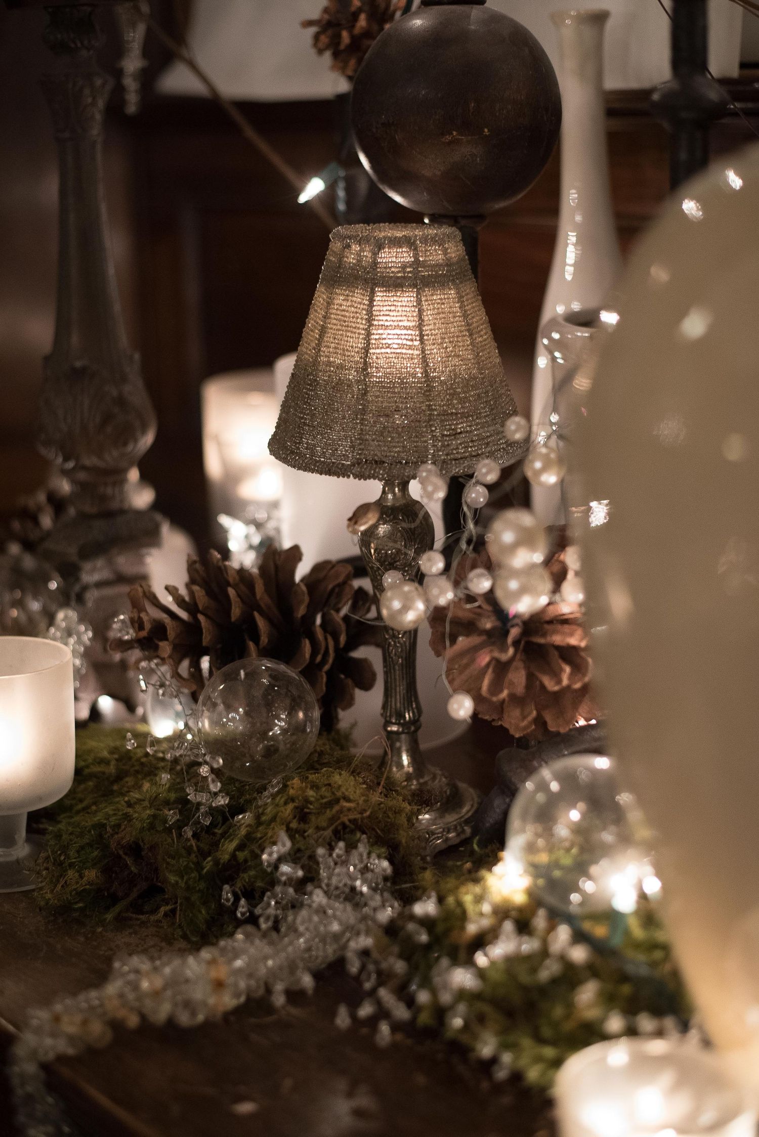 Cozy Christmas table decor with burlap lamp, pine cones, fairy lights, and glass ornaments in warm candlelight.