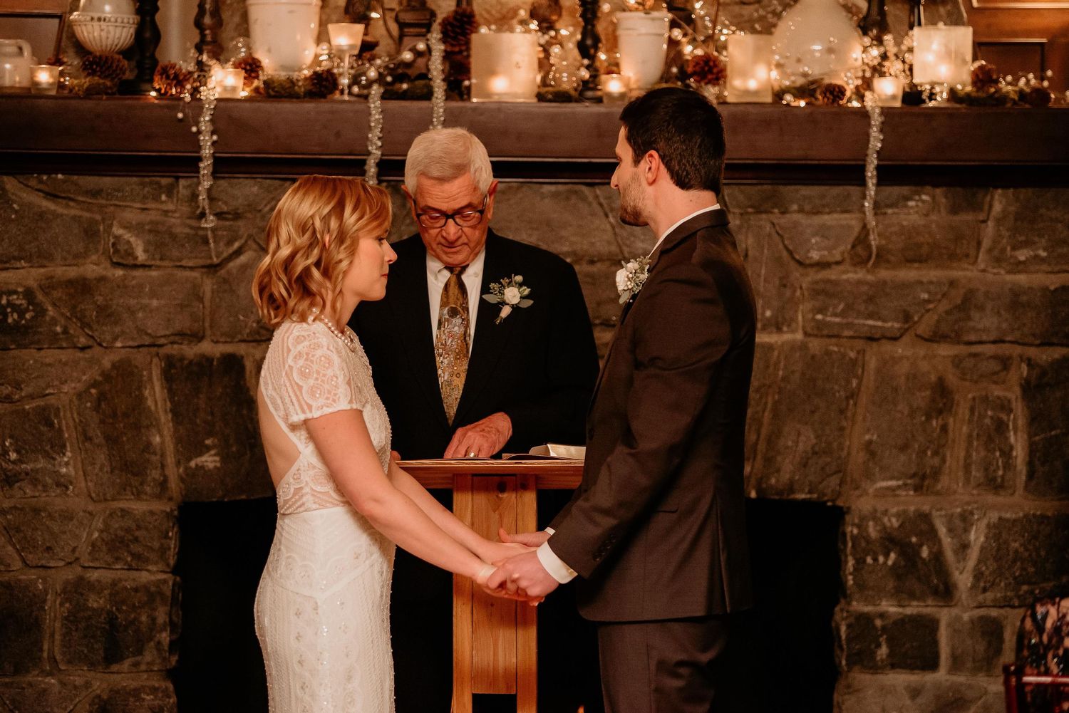 Bride and groom exchange vows before an officiant at a romantic indoor wedding ceremony by a stone fireplace.