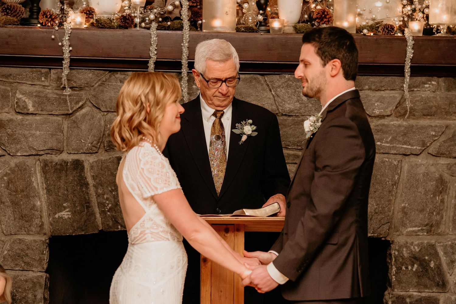 Bride and groom holding hands at indoor wedding ceremony before officiant at stone fireplace altar.