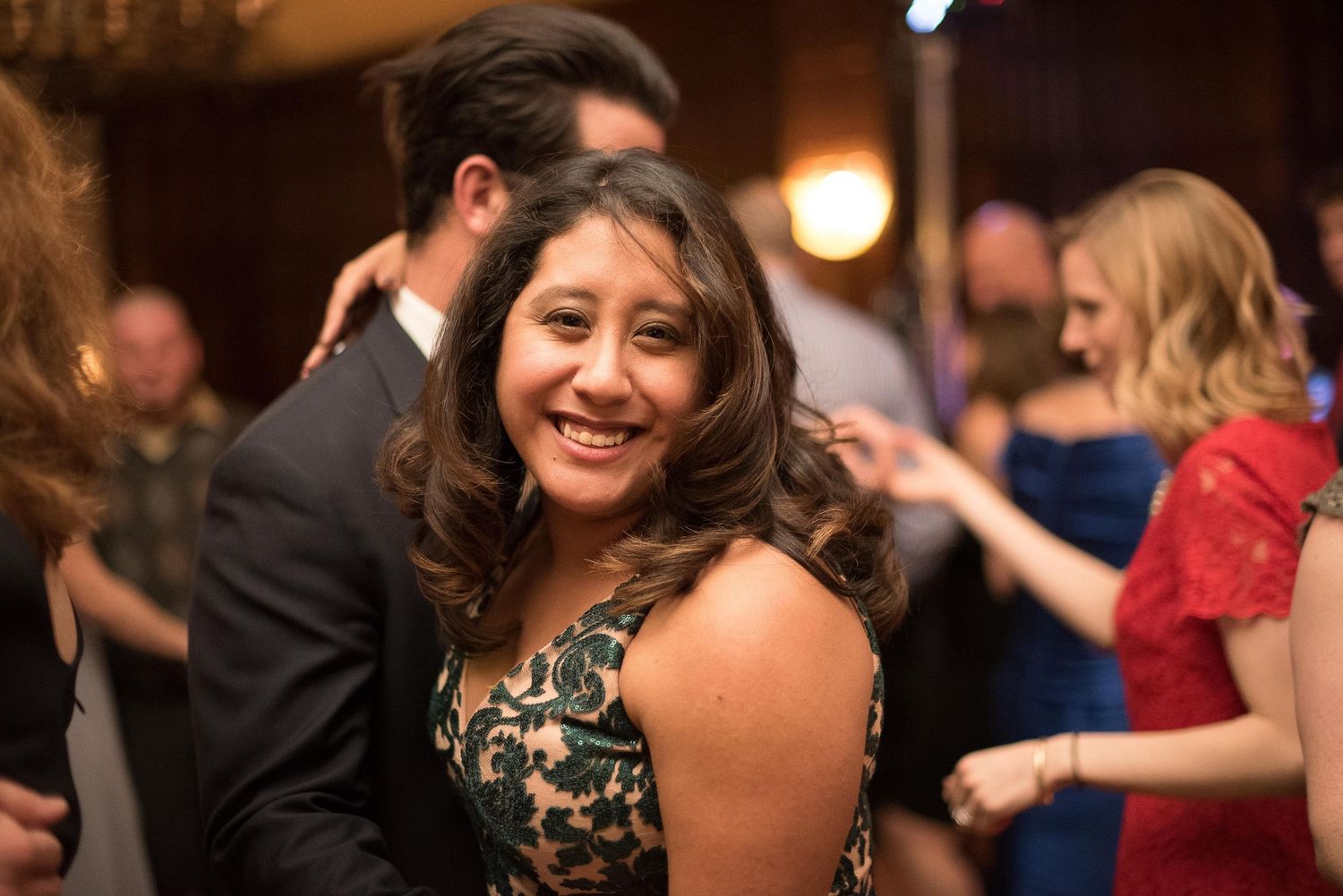 A woman in a floral dress smiles and dances at an elegant evening event with guests in formal attire.