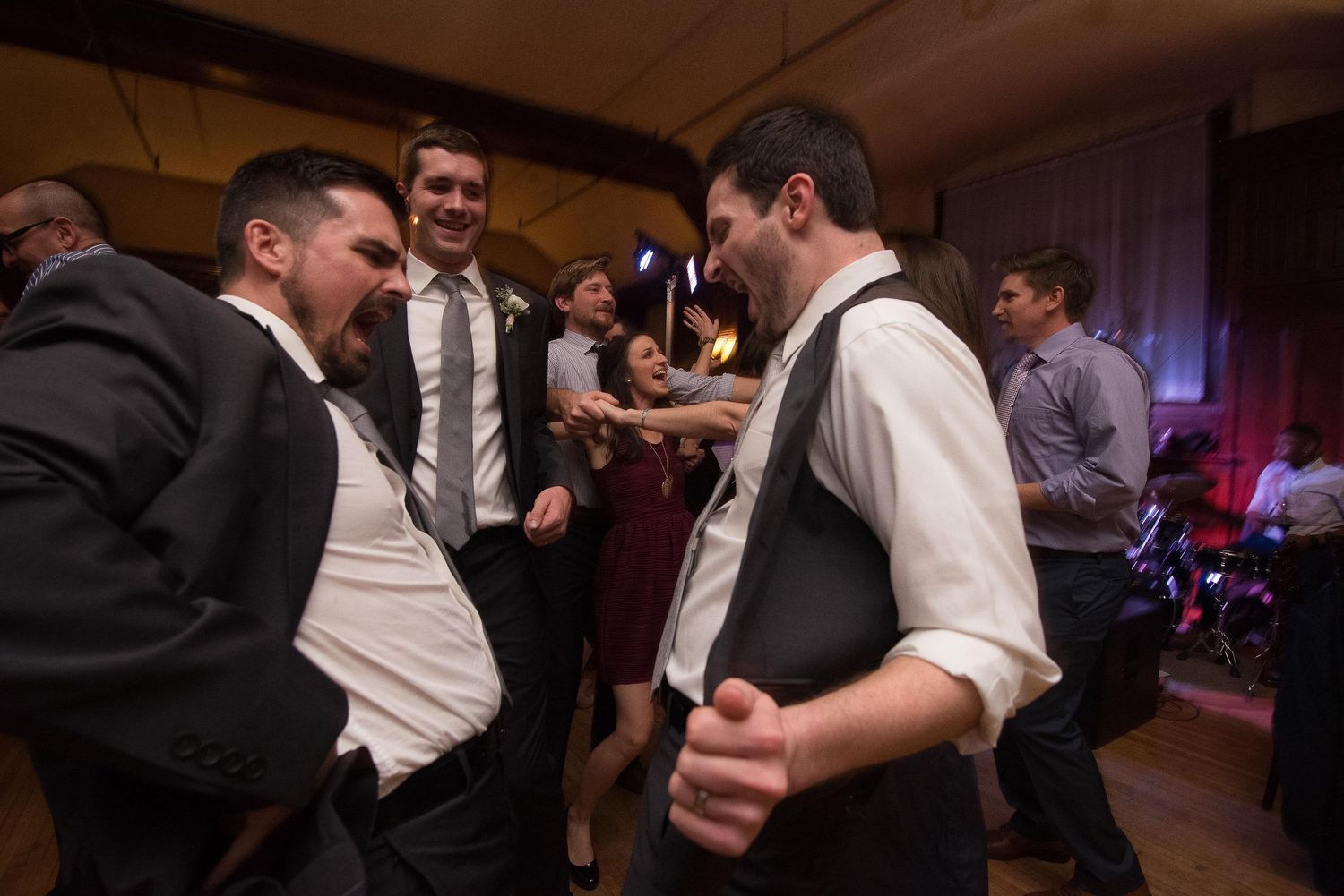 Men in formal attire laugh and dance together at a lively wedding reception celebration.