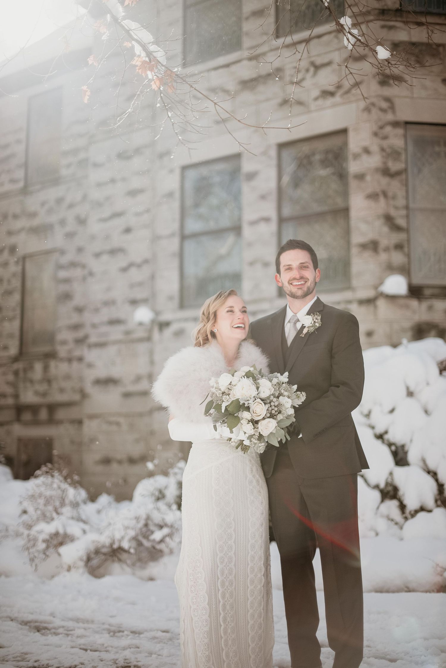 Bride and groom smile together in snow outside stone building, bride holding white bouquet wearing fur wrap.
