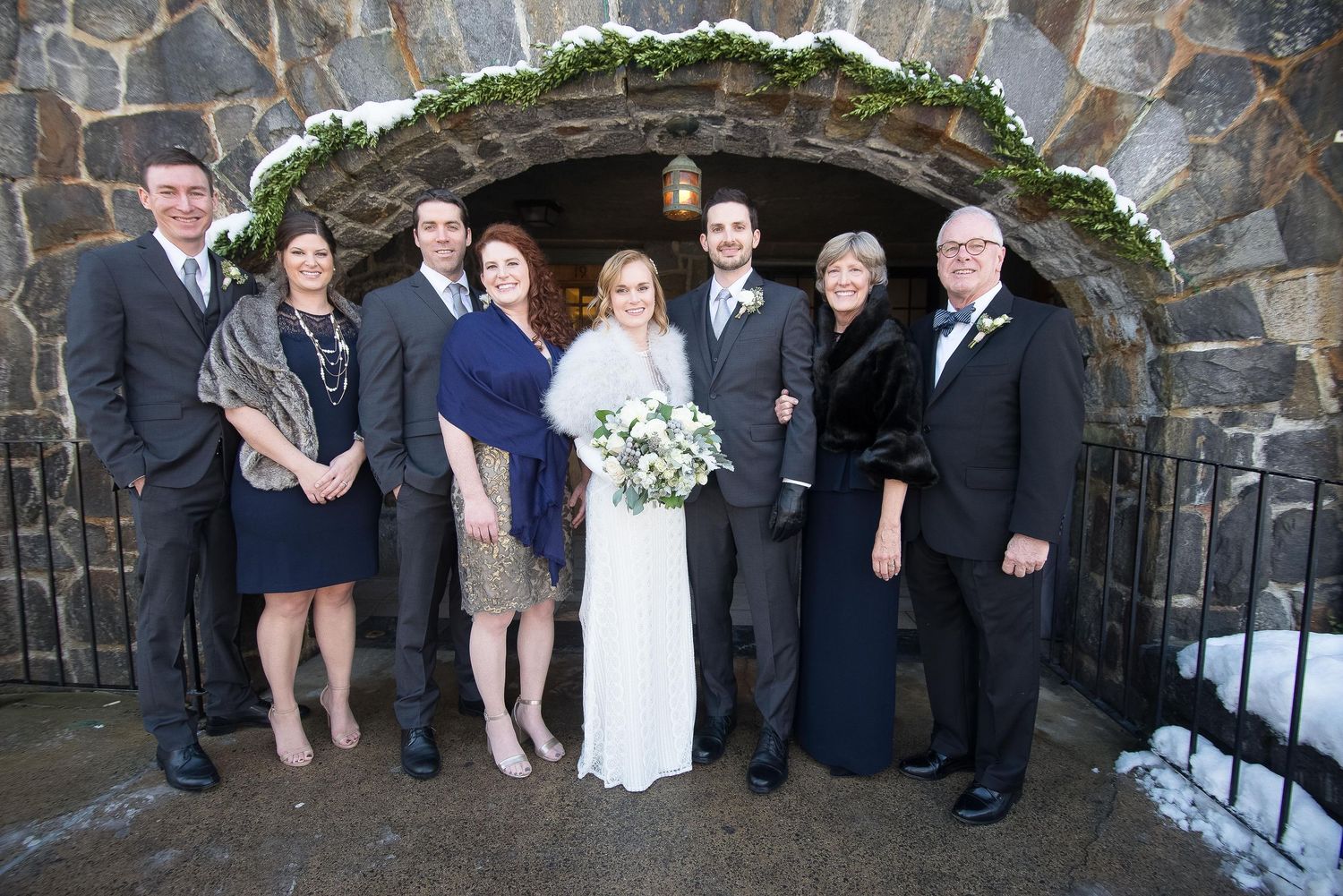 Wedding party poses in formal attire before a stone archway decorated with winter greenery in snowy setting.