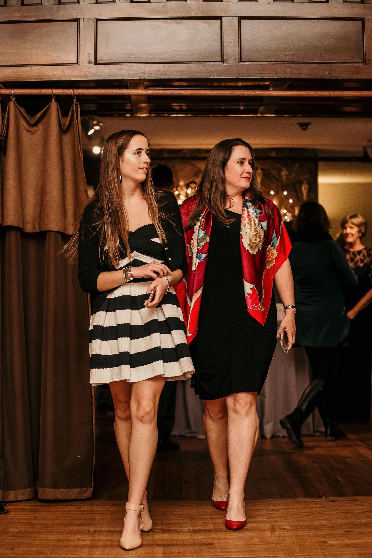 Two elegant women in black dresses walk together at an indoor event, one wearing a bold red floral scarf.