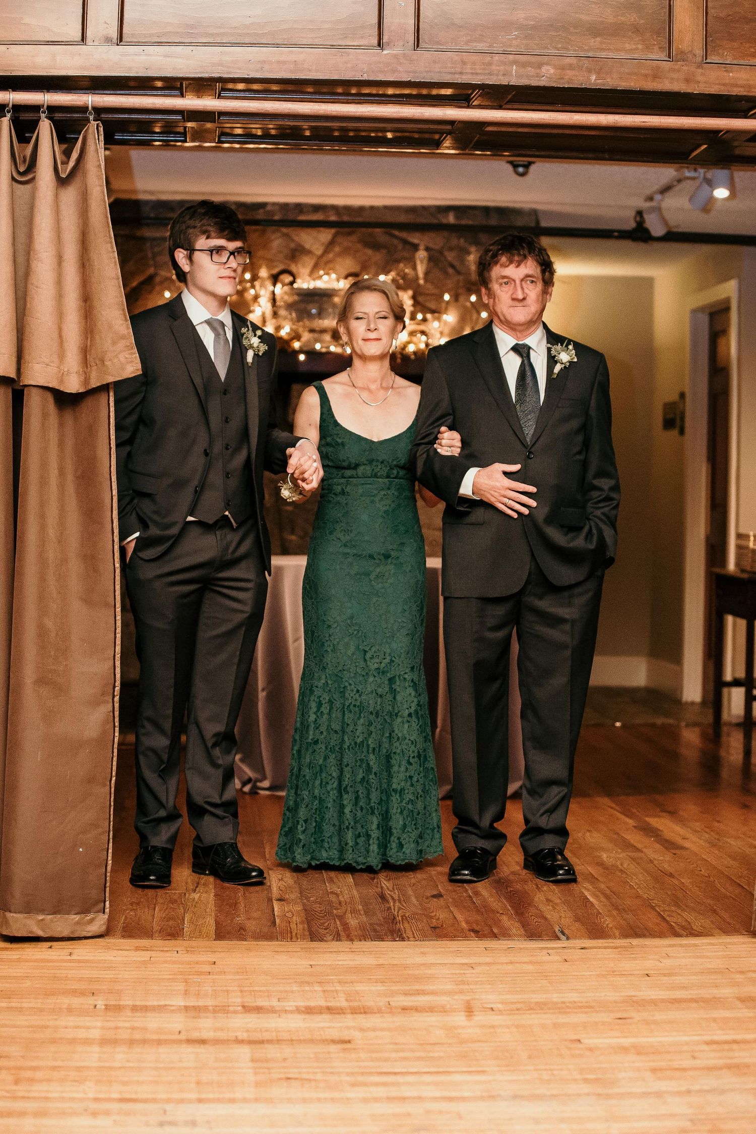 Woman in green lace gown escorted by two men in black suits at a wedding reception.