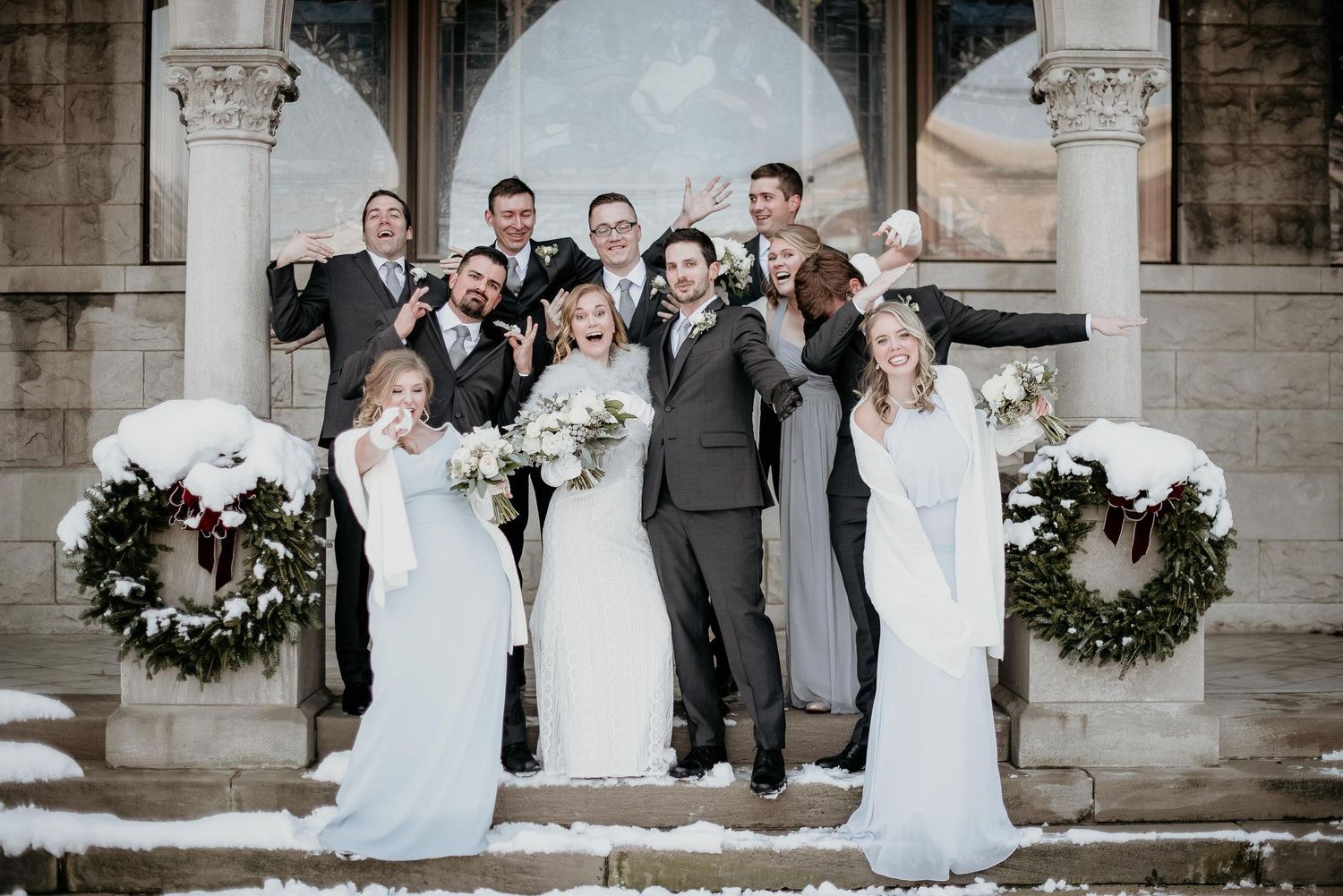 Winter wedding party poses on snowy steps, bridesmaids in white gowns, groomsmen in gray suits, with festive wreaths.