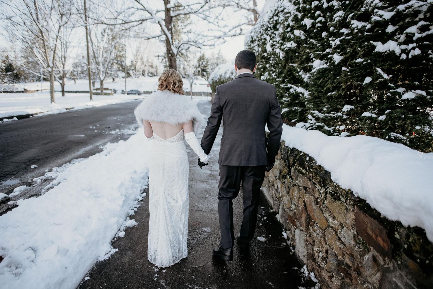 Bride and groom walking hand in hand along a snowy winter path, bride wearing elegant white gown with fur wrap.