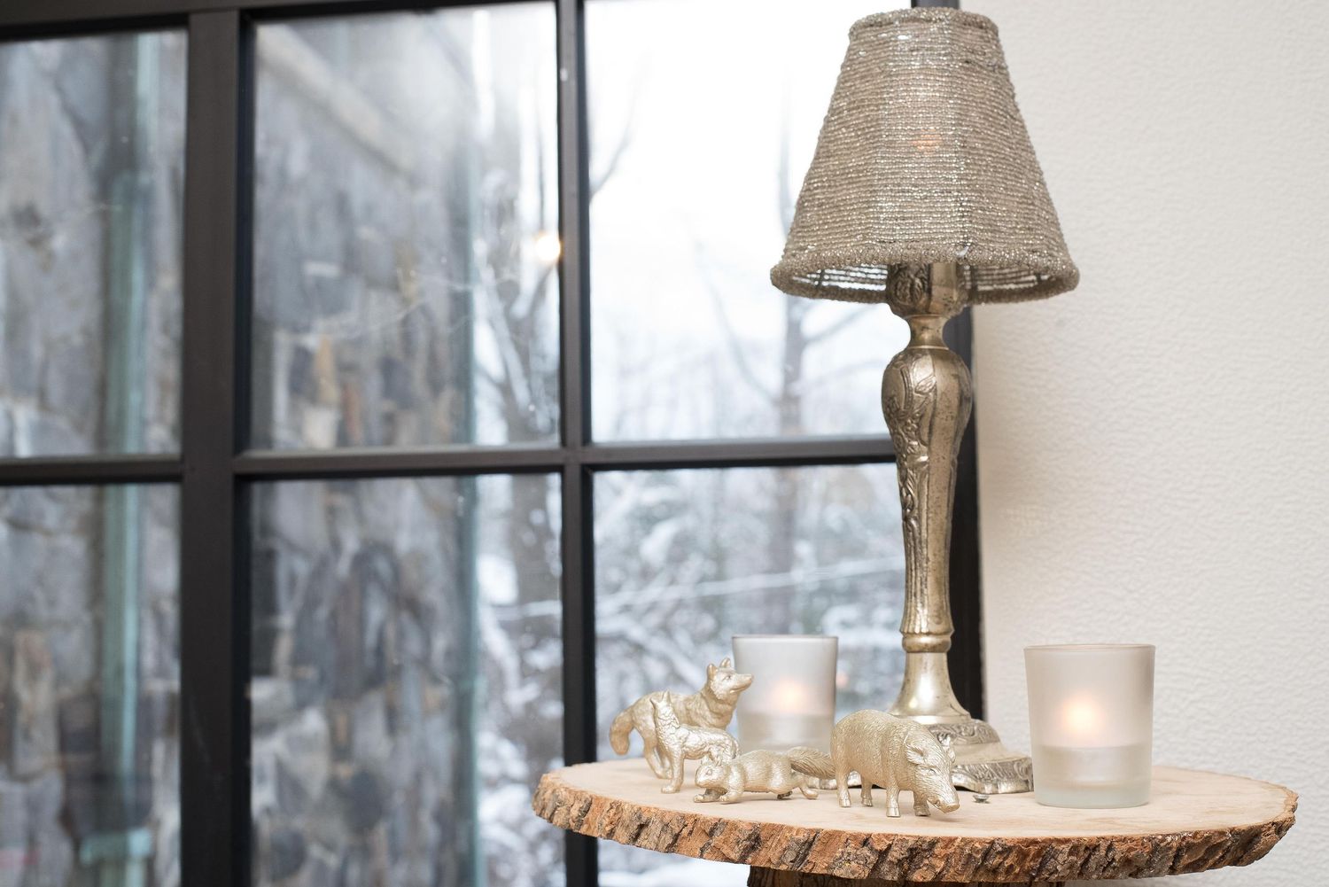 Silver lamp and glass candle holders on a rustic wood slice table by a snowy window.