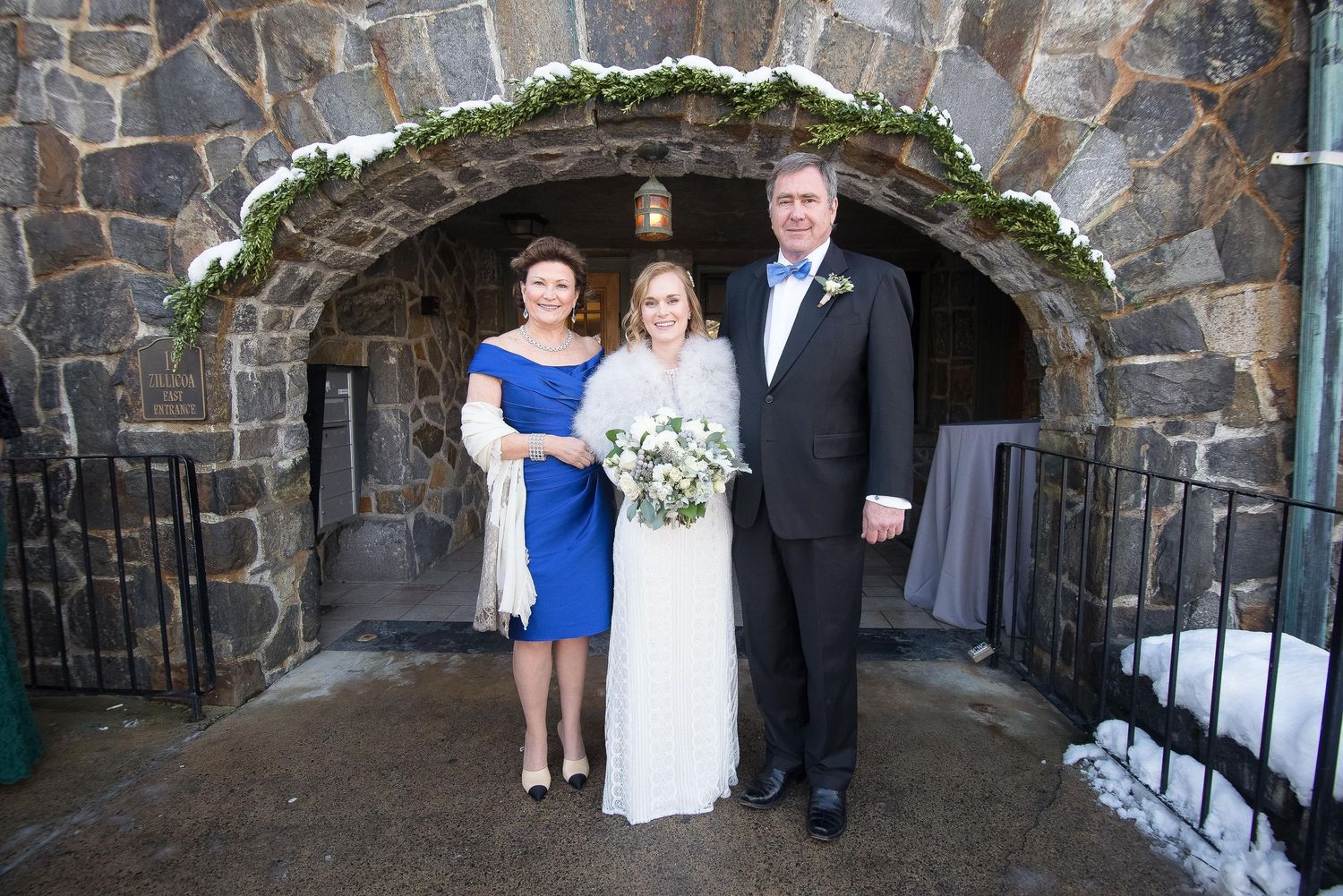 Bride in white gown poses with parents under stone arch decorated with winter greenery at elegant wedding venue.