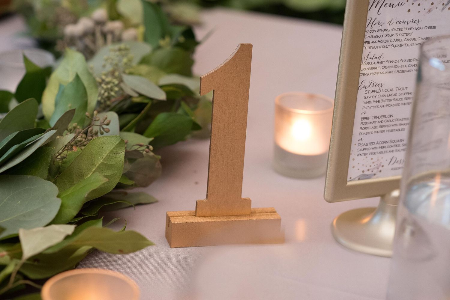 Gold number one table marker beside greenery, candles, and a framed menu at an elegant wedding reception.