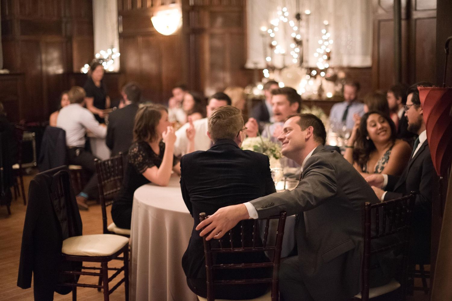 Elegant wedding reception guests seated at round tables in a warmly lit venue with string lights and wood paneling.