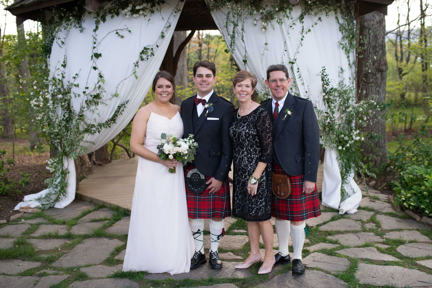 Wedding party of four posing under floral gazebo, two men in red kilts flanking bride in white gown and woman in black dress.