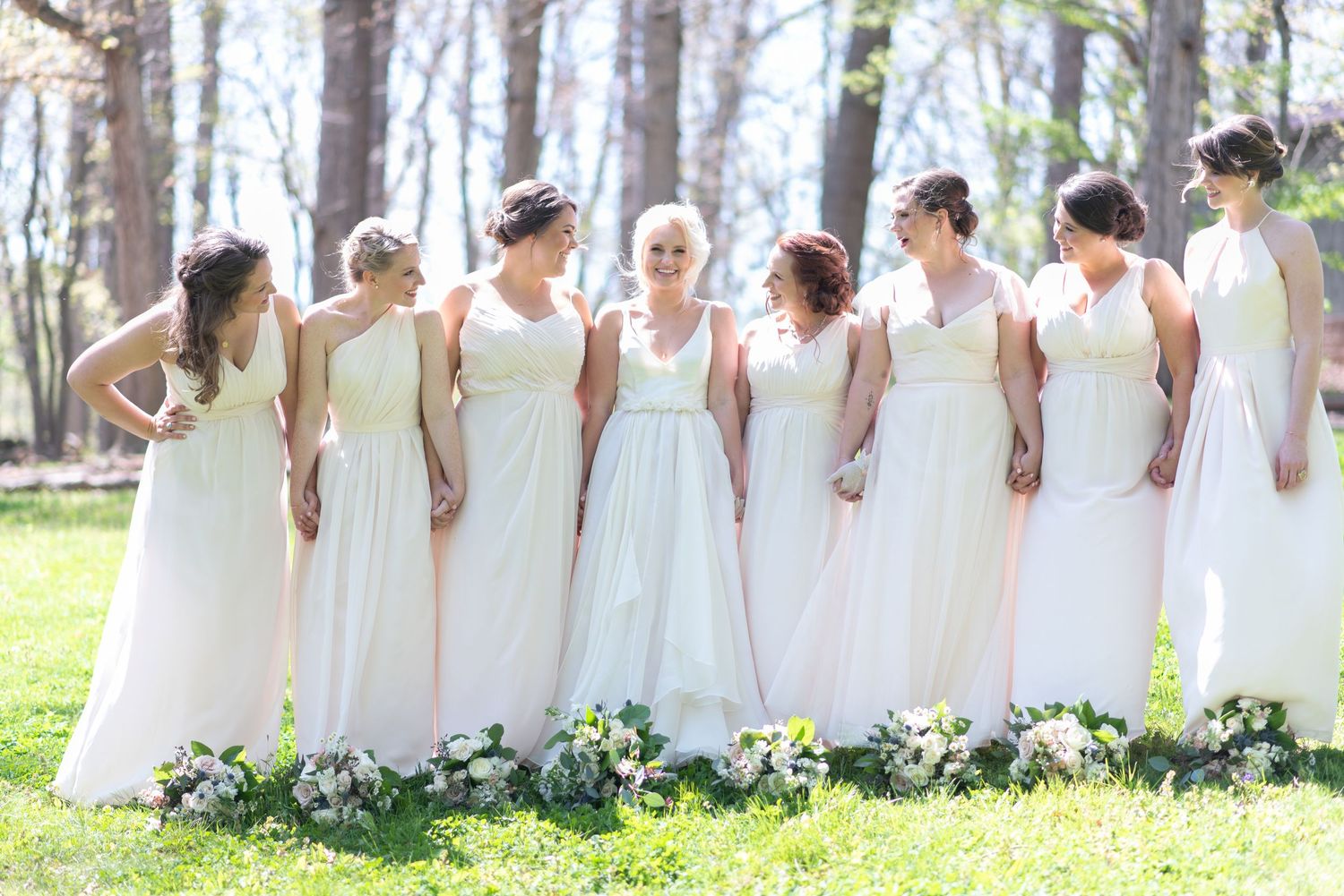 Bride stands with seven bridesmaids in matching ivory gowns on green lawn in spring.