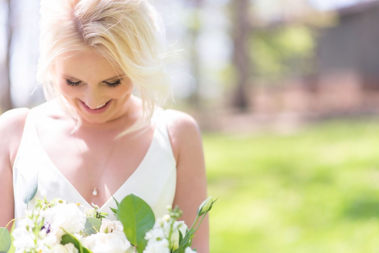 Radiant blonde bride in V-neck gown smiles down at white wedding bouquet outdoors.
