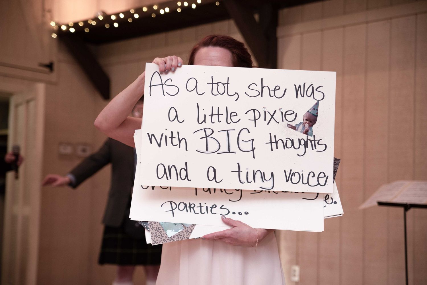 A flower girl holds a sign with a sweet poem about the bride at an indoor wedding reception.