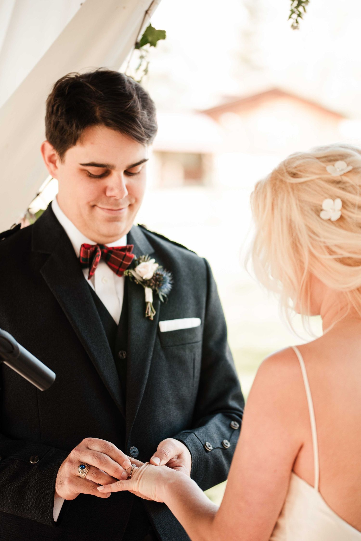 A groom in a black suit with tartan bow tie places a ring on his bride's finger during the ceremony.