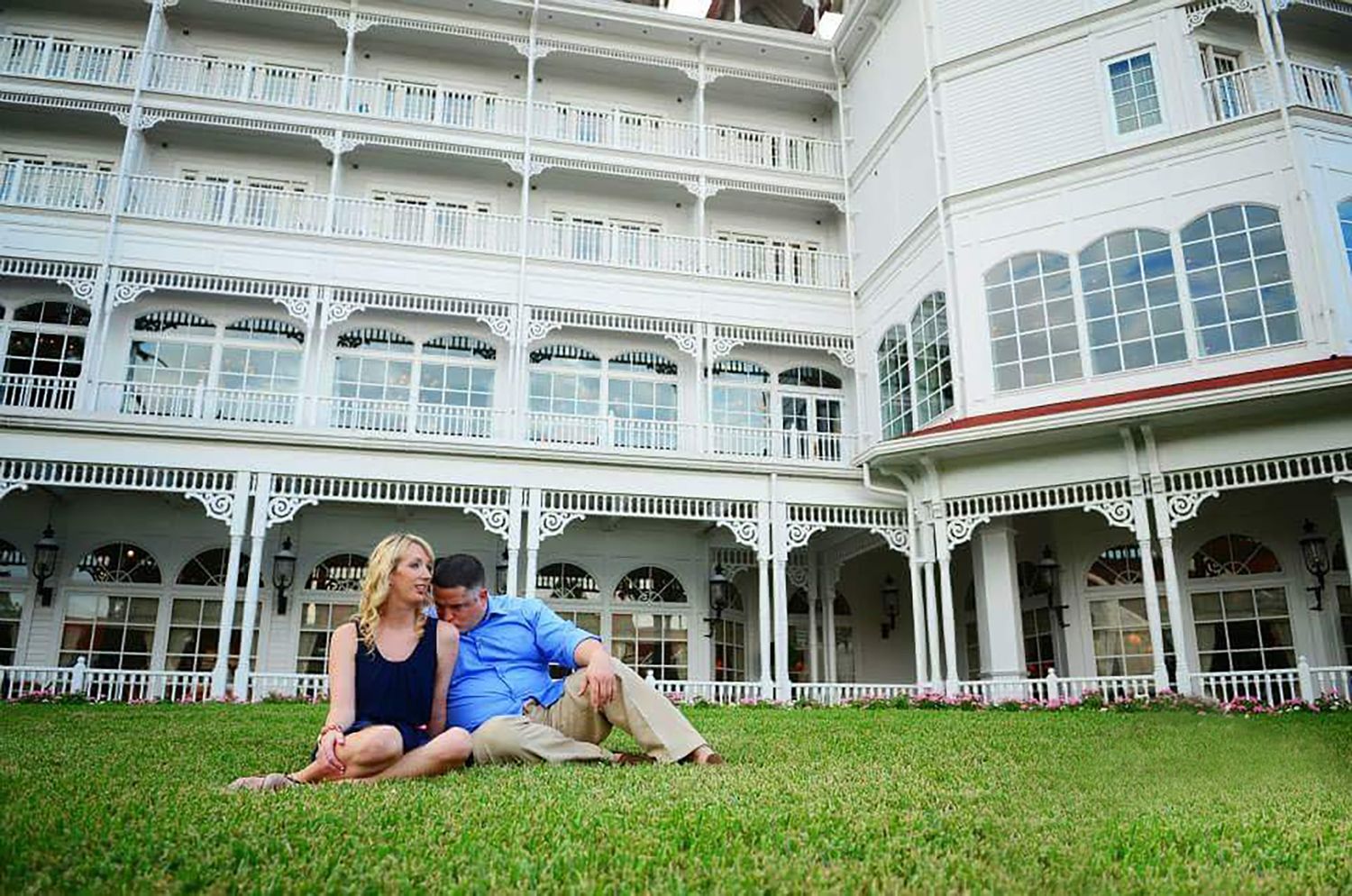 Couple sitting together on green lawn in front of elegant white Victorian hotel building.