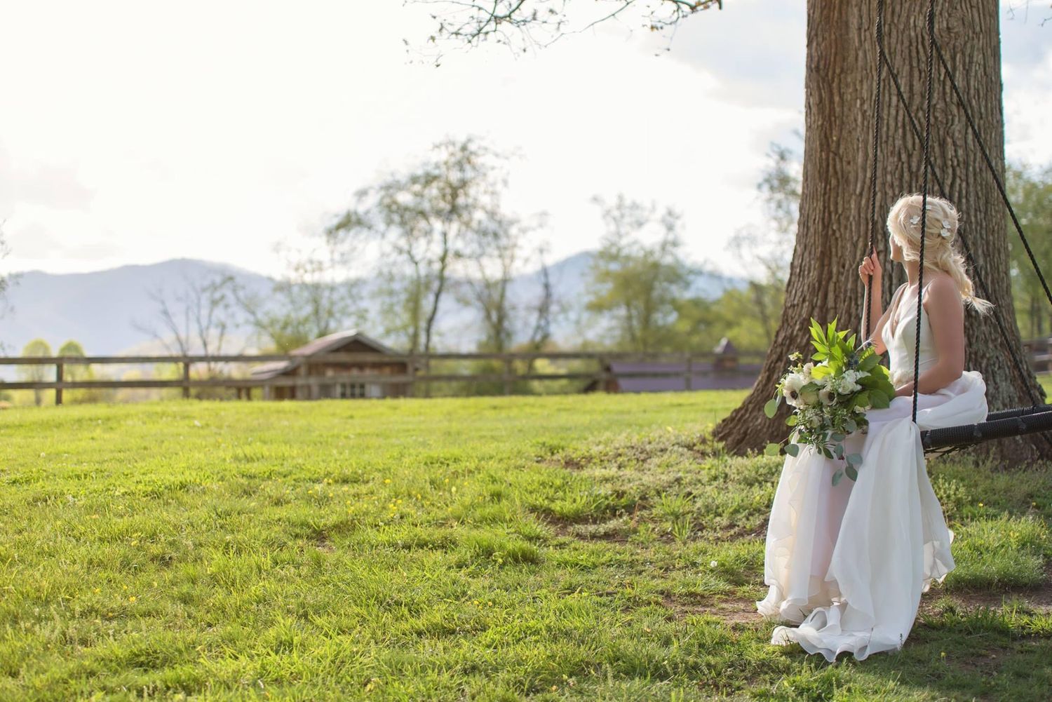 A bride in a white gown leans against a large tree in a lush green farm setting with mountain views.