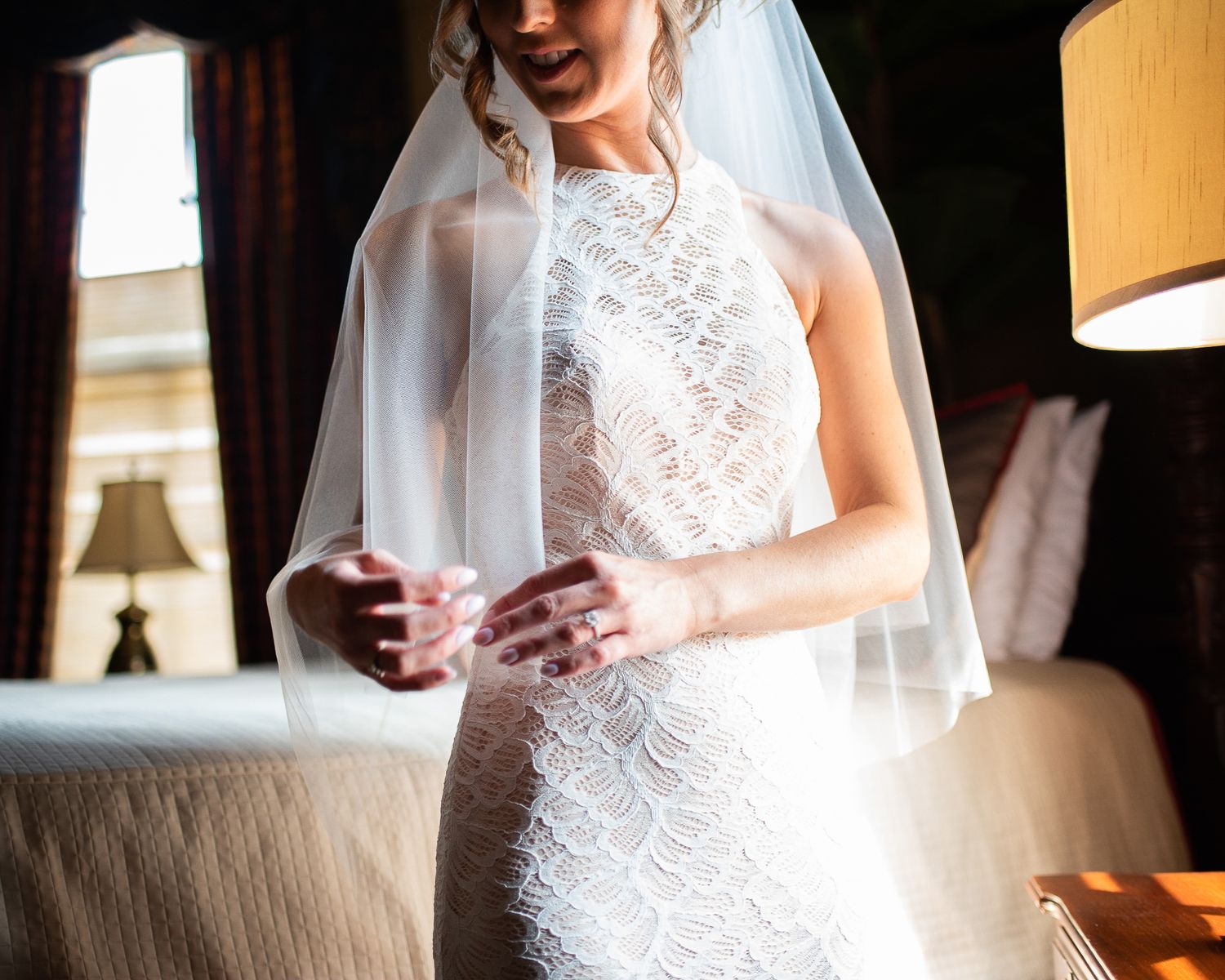 Bride in a halter-neck lace wedding gown and veil stands in a softly lit hotel room.