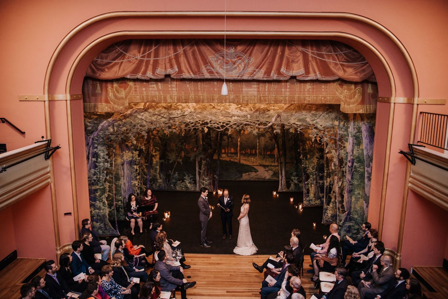 Aerial view of a wedding ceremony on a theater stage with ornate pink arch and seated guests.