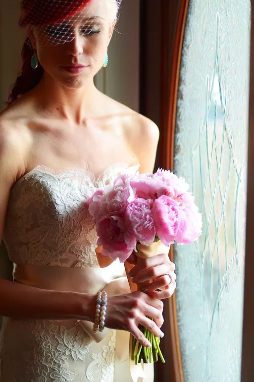 Bride in lace strapless gown holding pink peony bouquet with birdcage veil by window.