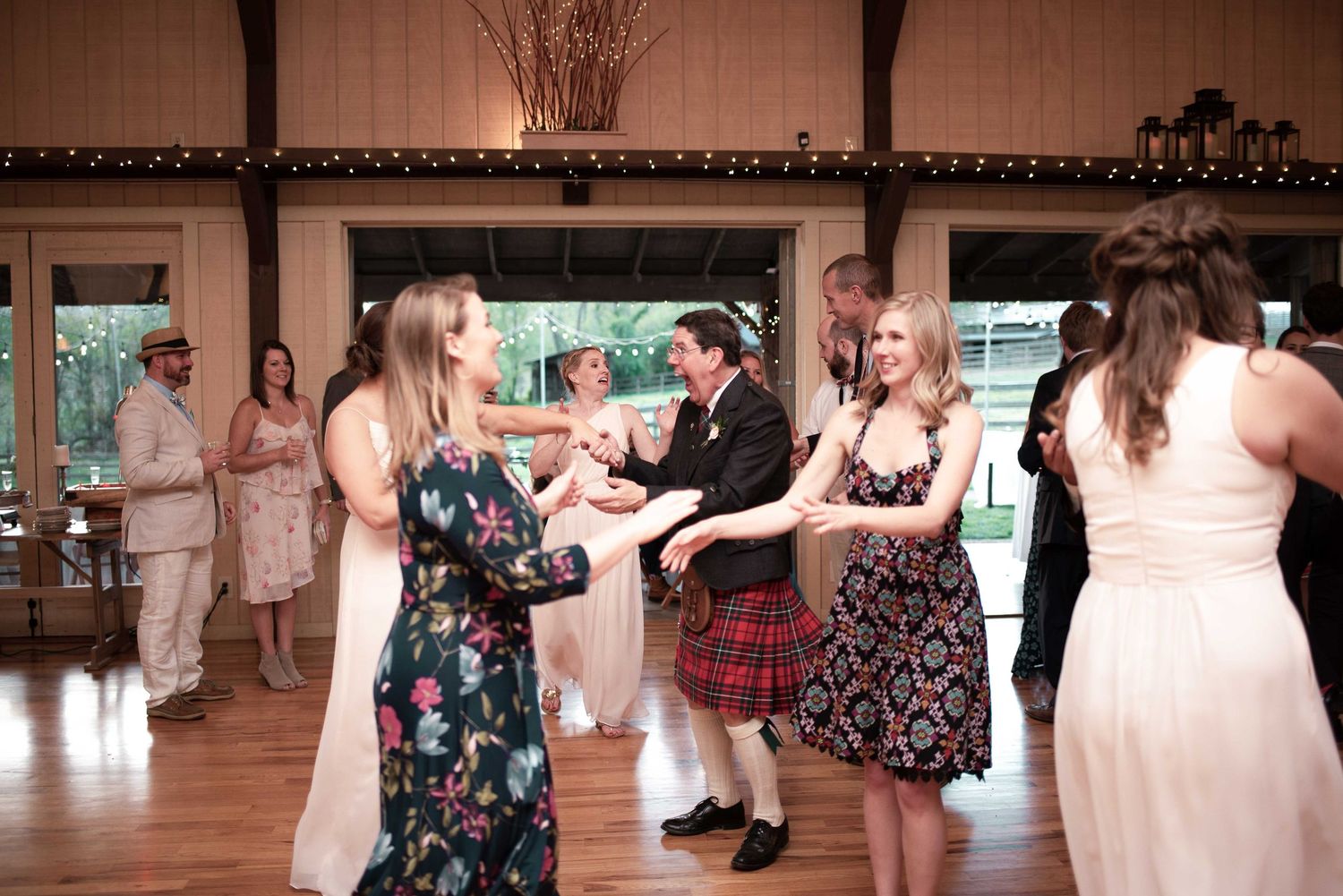 Guests dancing at a wedding reception, including a man in a red tartan kilt, in a rustic barn venue.