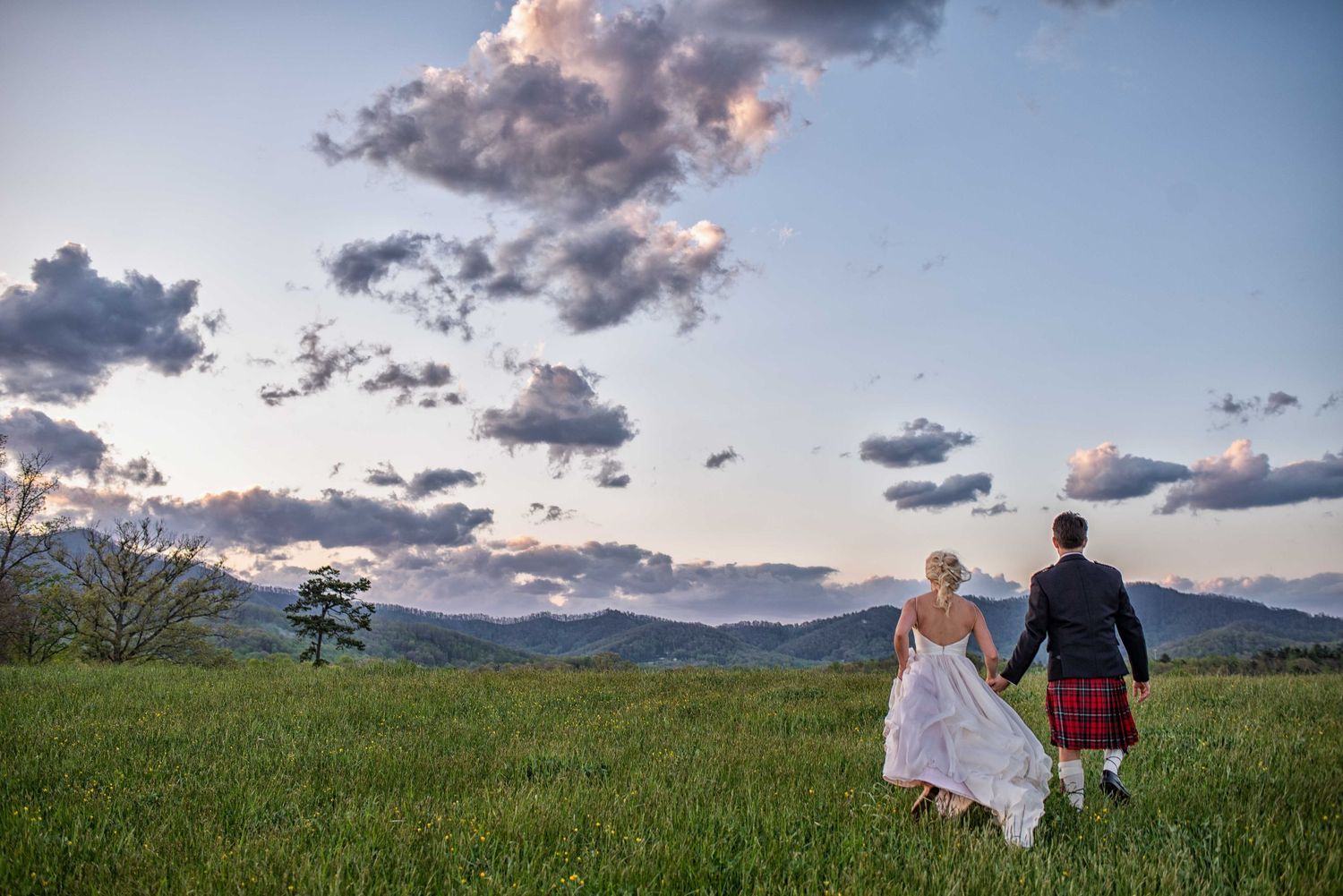 Bride and groom in Scottish kilt walking hand-in-hand through green meadow with mountain sunset backdrop.