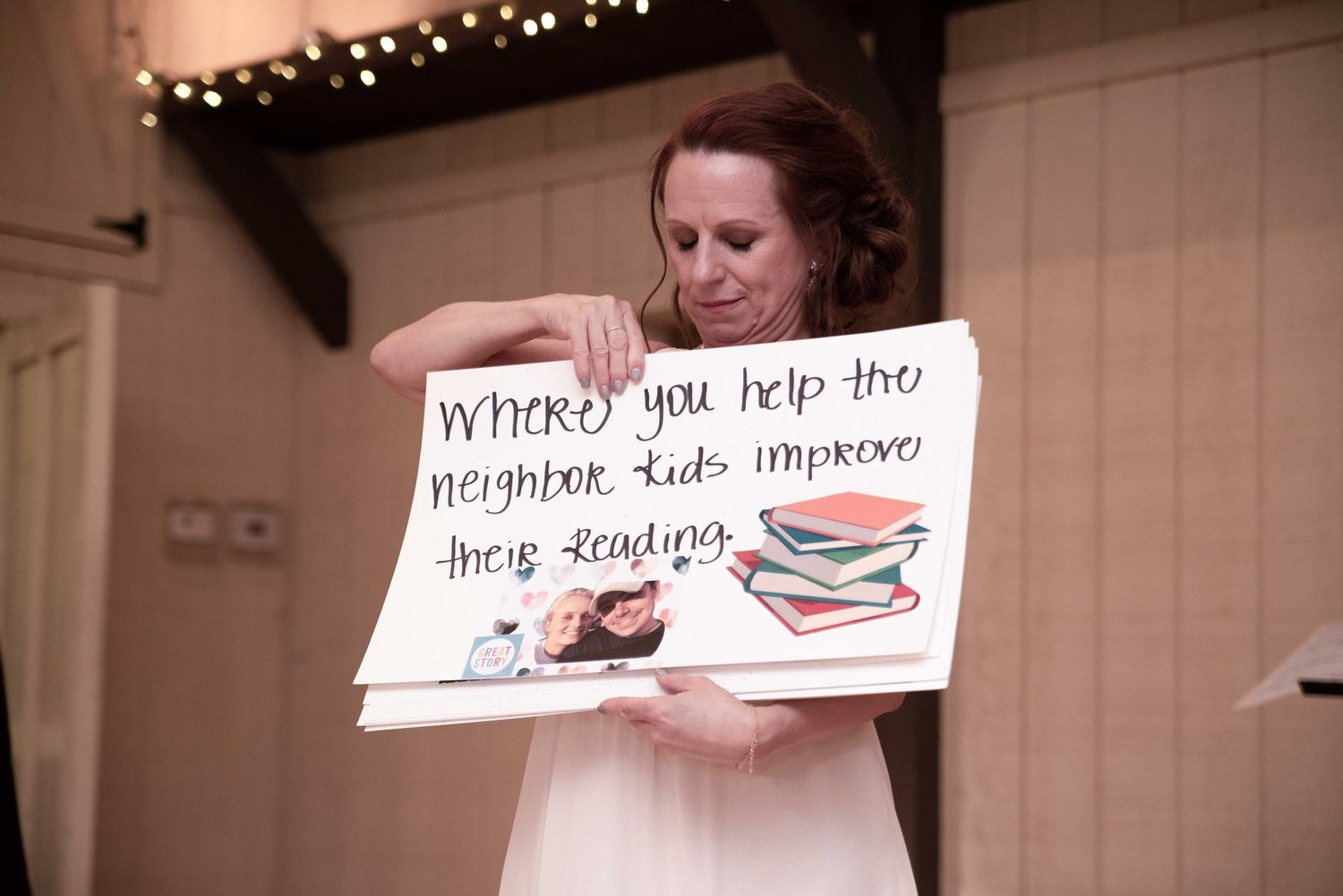 Woman holding a handmade sign about helping neighbor kids improve their reading at an indoor event.