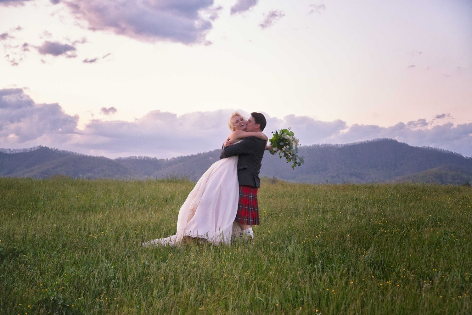 Couple embracing in green meadow at sunset, groom in red tartan kilt, bride in white gown holding bouquet, mountains behind.