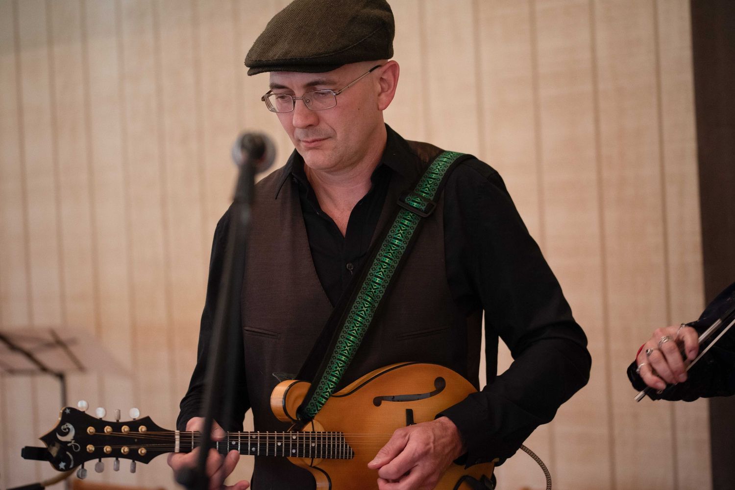 Musician playing mandolin with green strap, wearing black shirt and flat cap, performing at an indoor event.