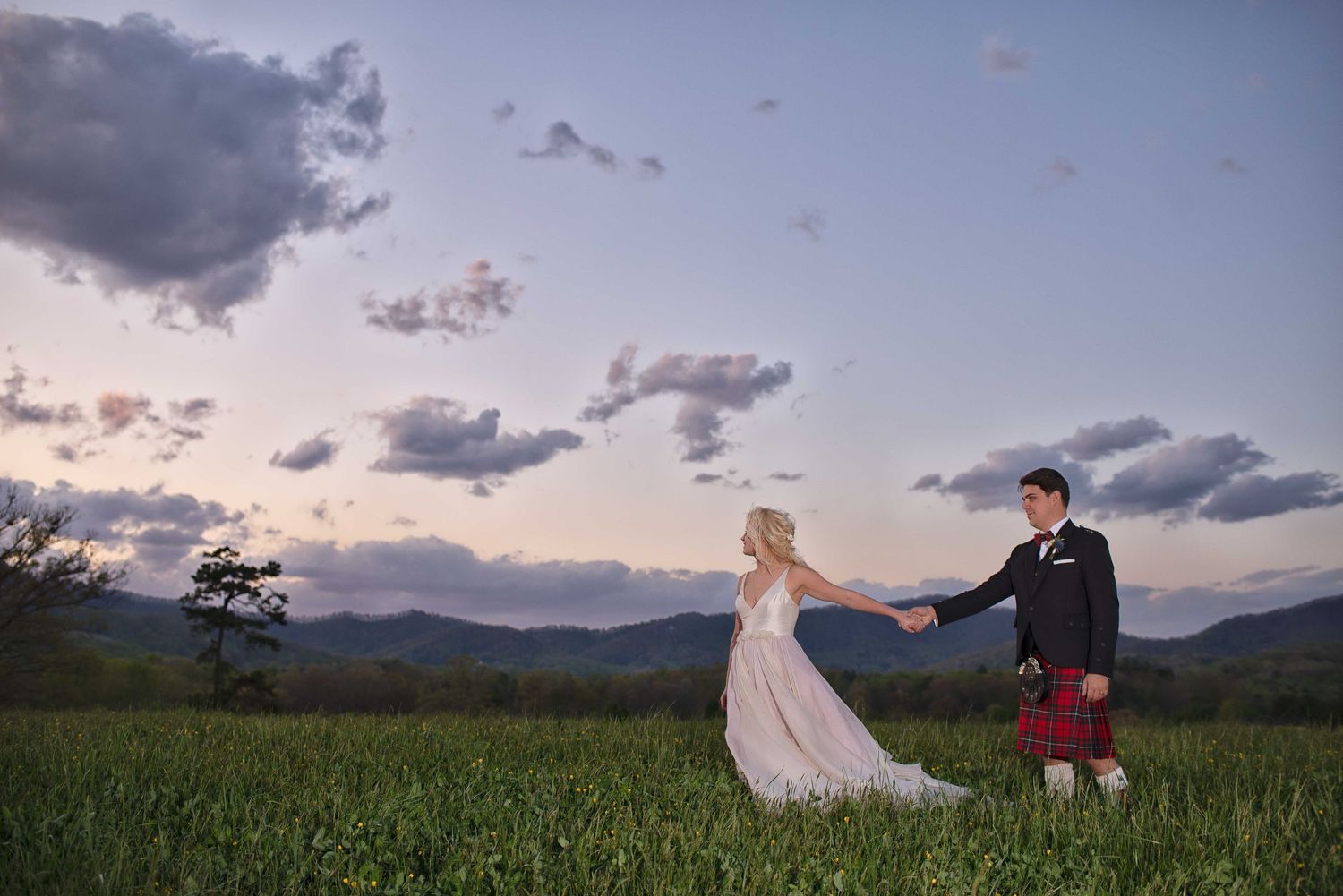 Bride and groom holding hands in a lush green field at sunset with mountain views, groom wearing a traditional Scottish kilt.