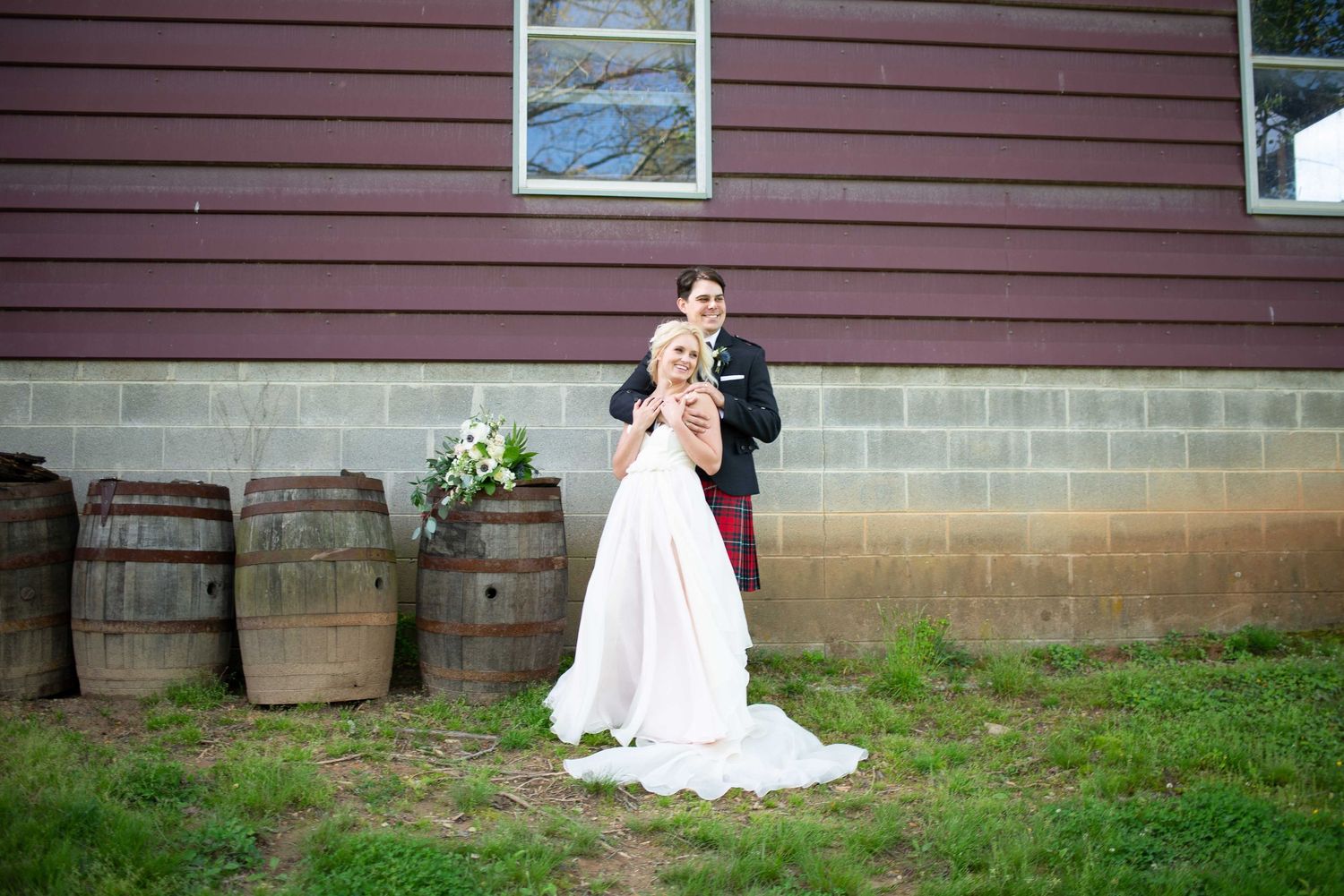 Bride and groom posing outdoors near rustic wooden barrels beside a red barn on their wedding day.