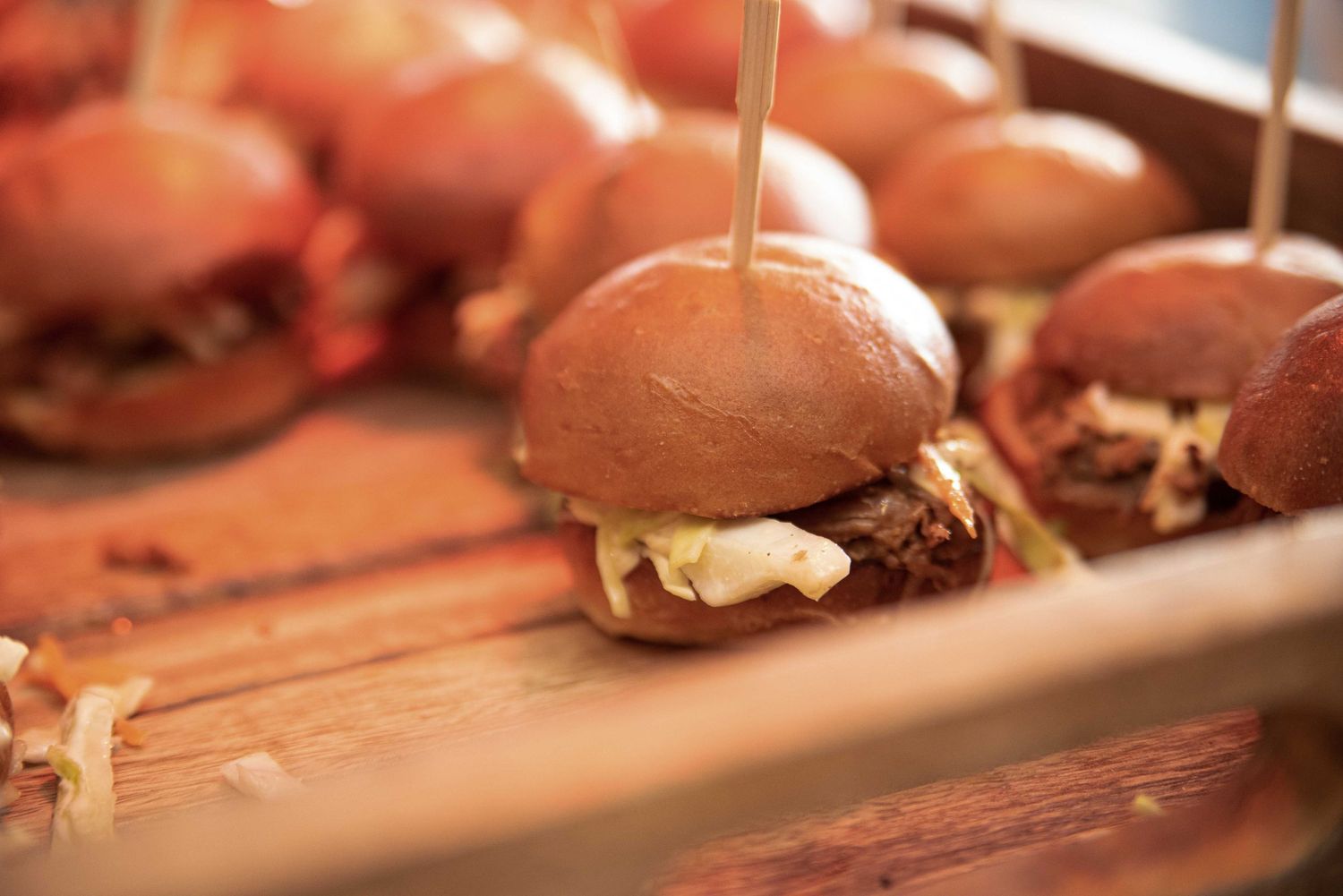 Close-up of gourmet mini sliders with melted cheese on a rustic wooden serving board at a catered event.