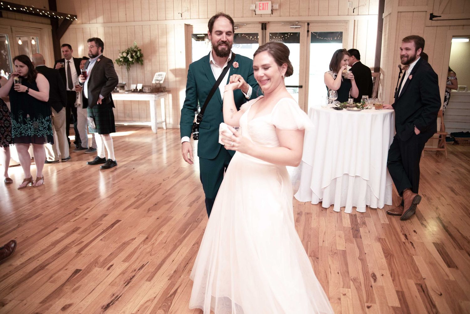 Bride and groom share first dance on wooden floor at wedding reception with guests in background.