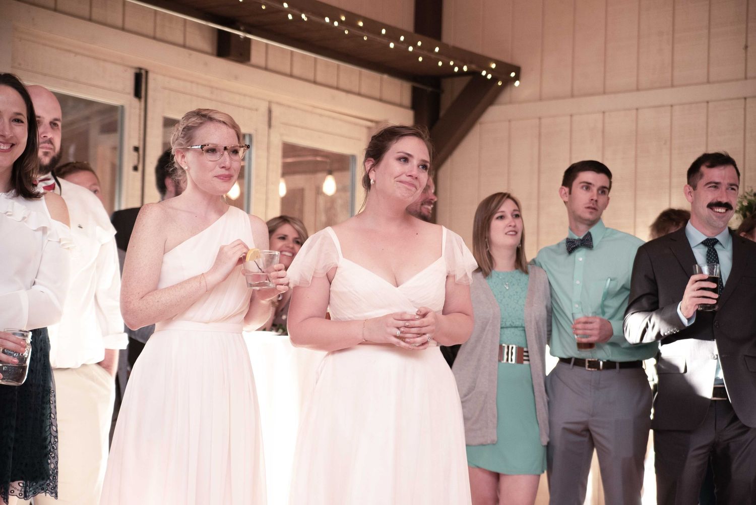 Bridesmaids in white and mint dresses stand together at a rustic barn wedding reception, holding drinks during toasts.