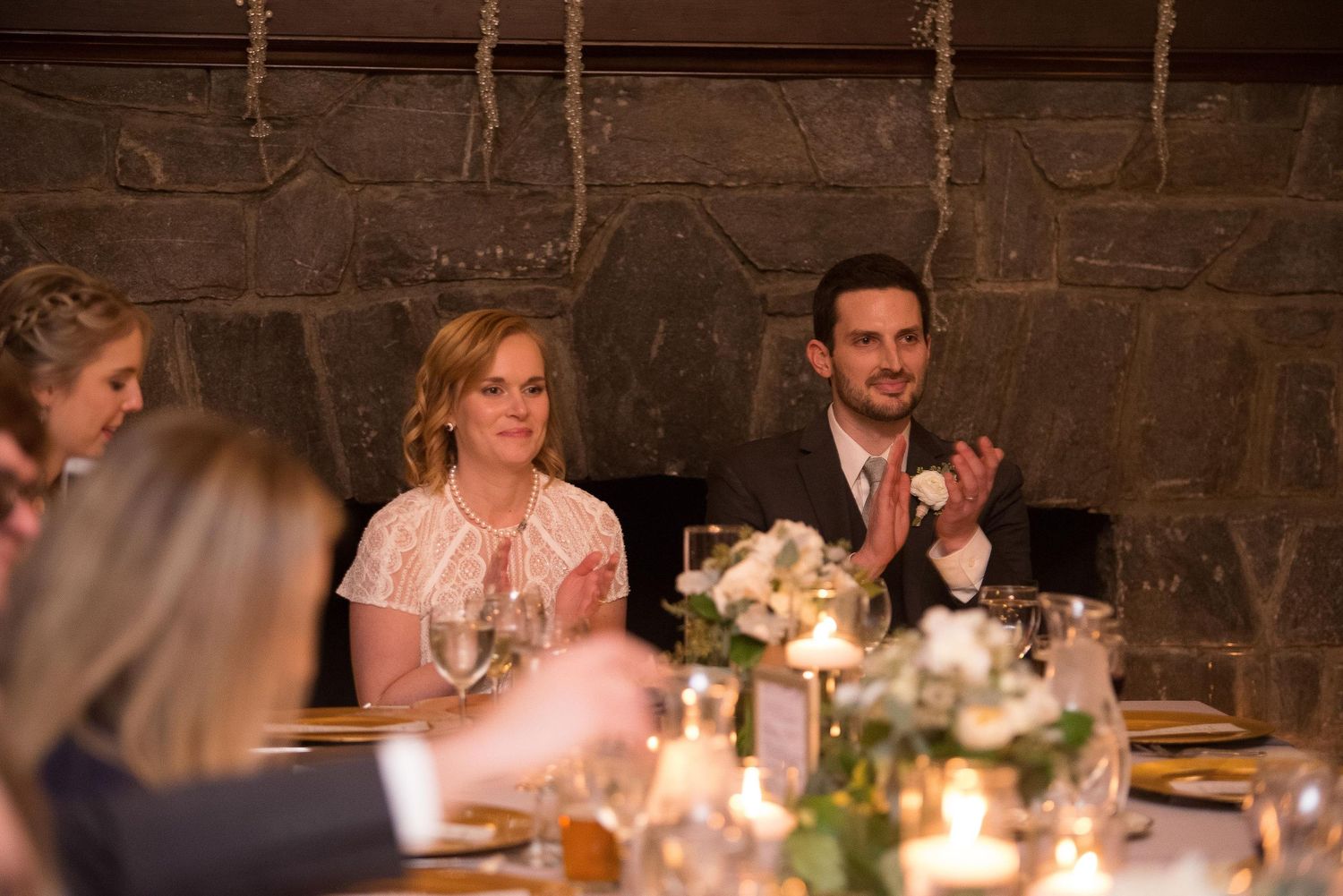 Couple in formal attire clapping at a candlelit wedding reception dinner table with floral centerpieces.