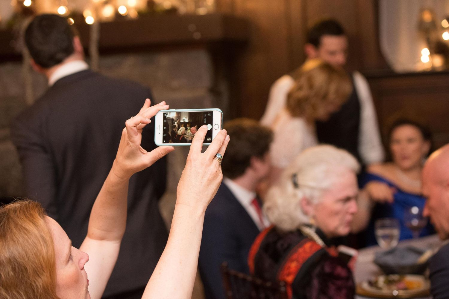 Guest captures wedding reception moment on smartphone, with bride and groom visible in warm, candlelit venue background.