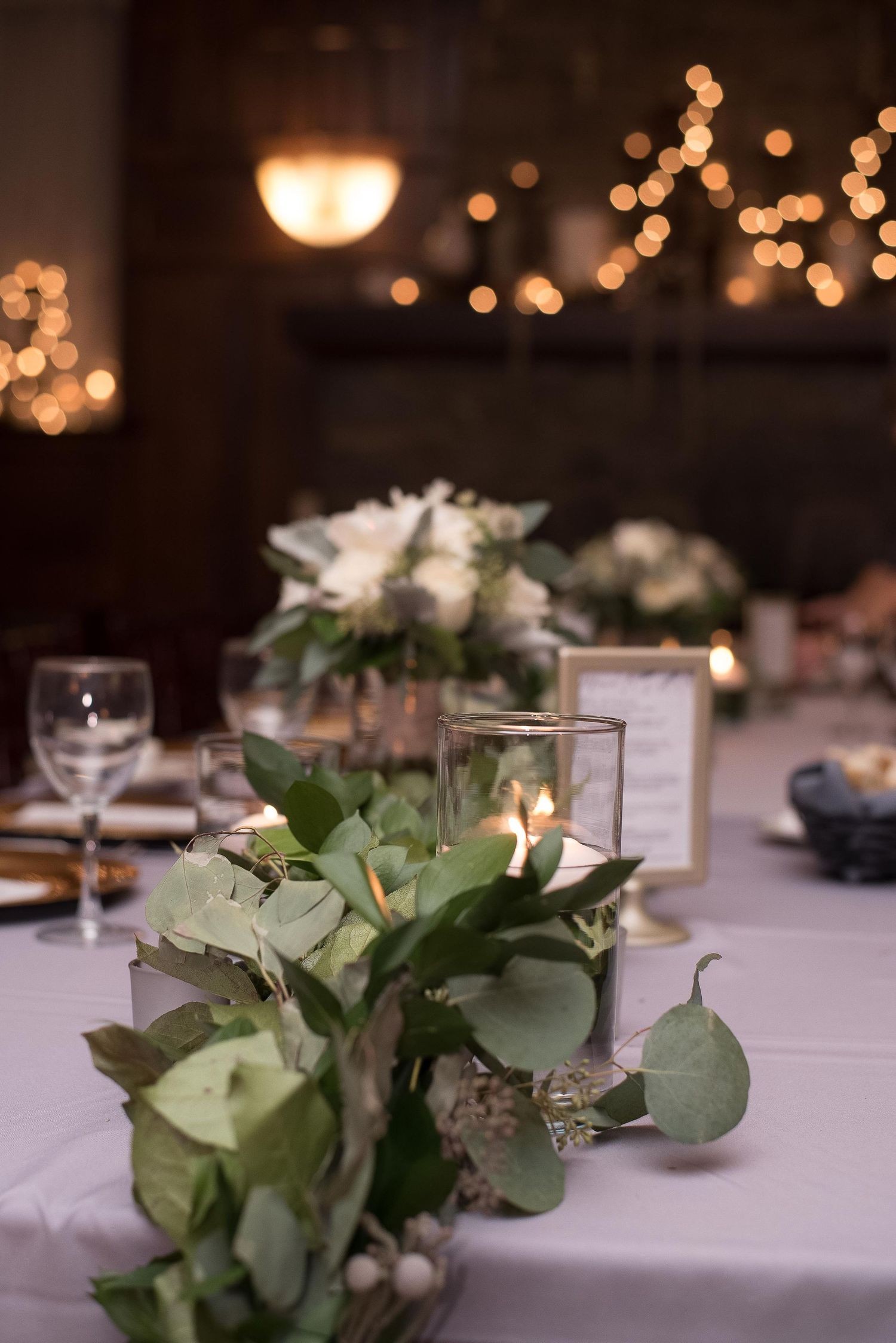 Elegant wedding reception table with eucalyptus greenery, candles in glass hurricanes, and soft bokeh string lights in background.